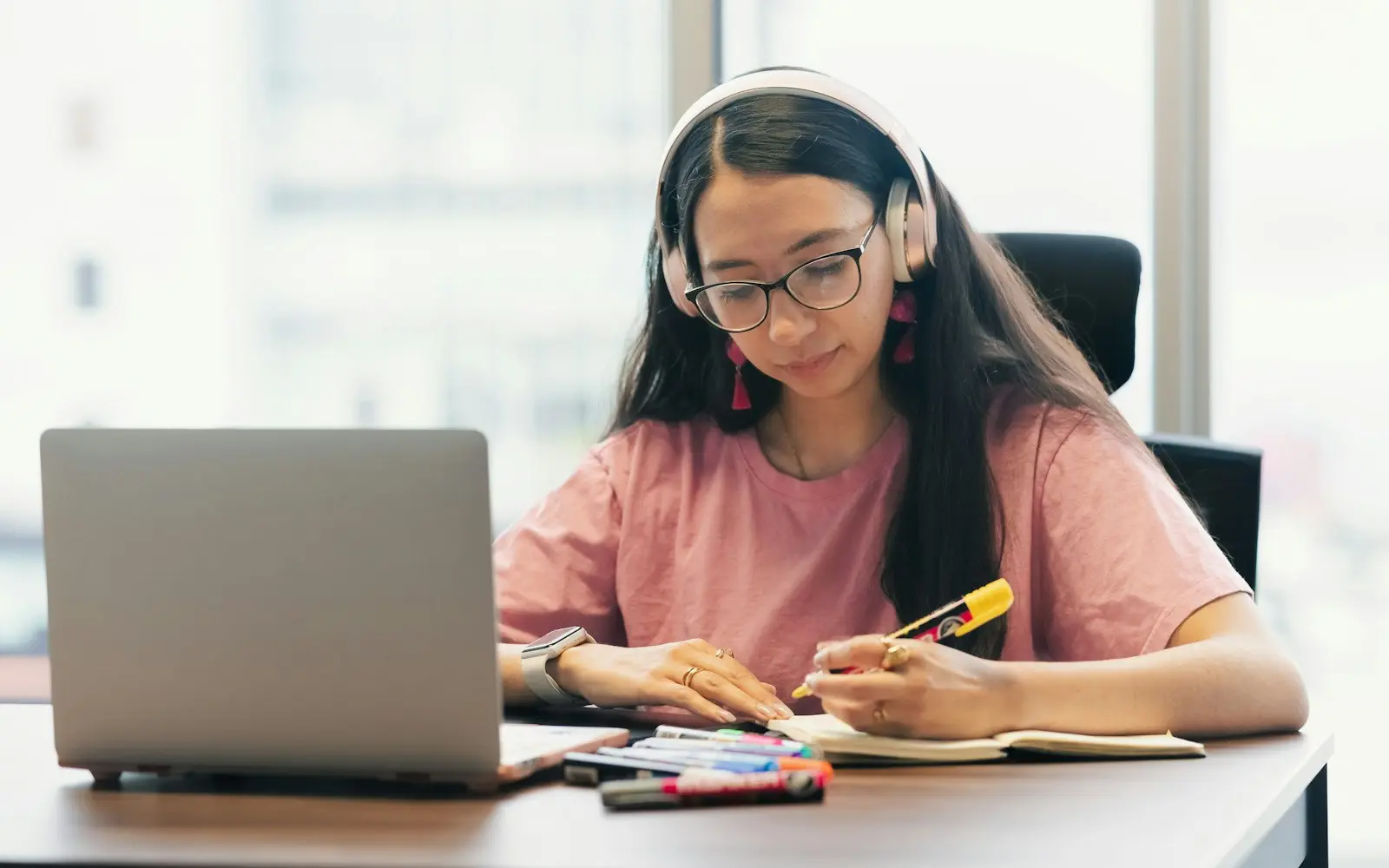 Young woman in headphones studying at a bright desk with a laptop and notebook